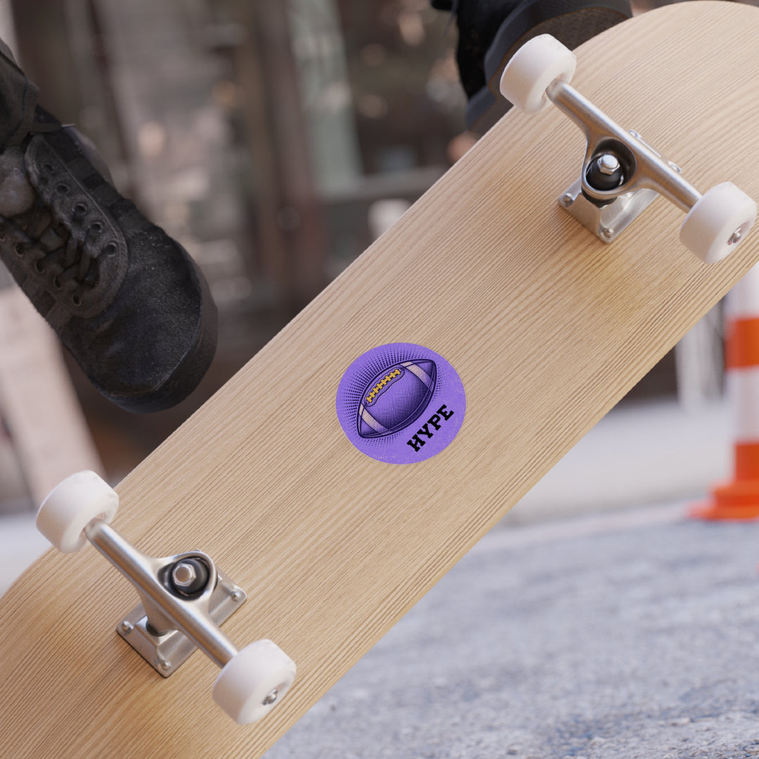A close-up of a skateboard deck with a purple circular sticker that reads &