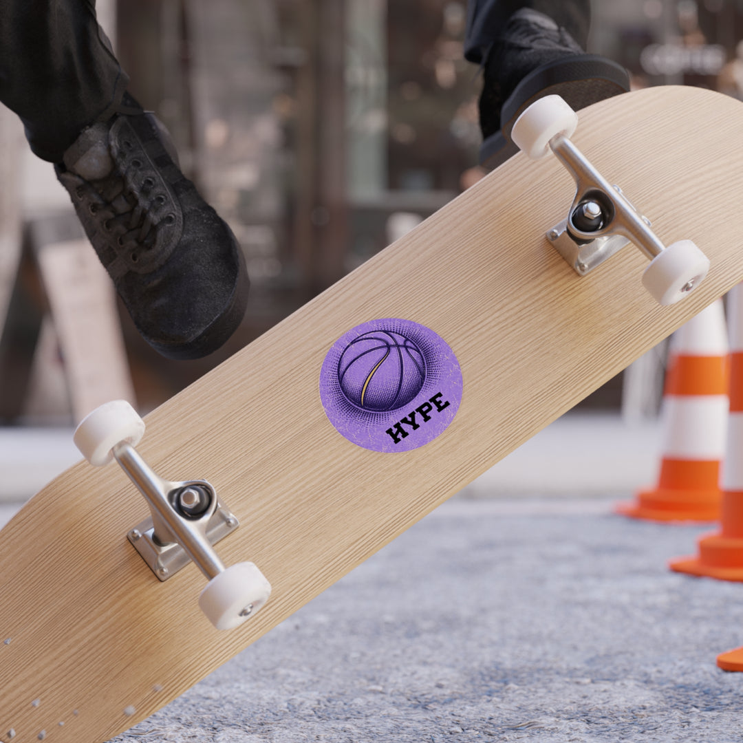 A close-up of a skateboard deck with a purple circular sticker featuring a basketball graphic and the text &