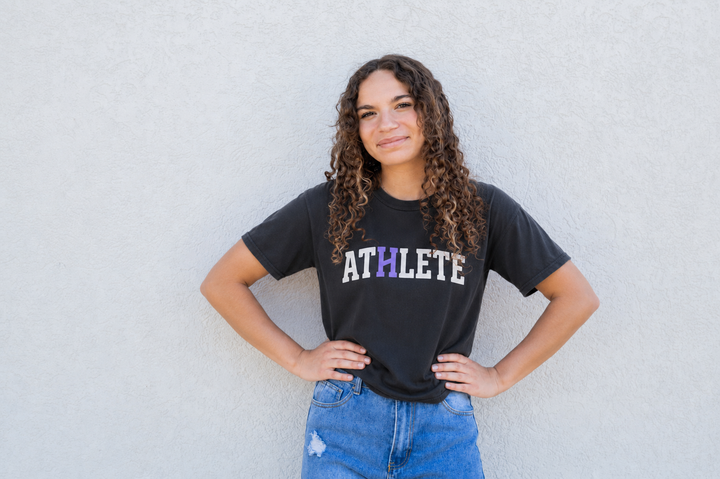 A young woman with curly brown hair stands against a light gray textured wall, wearing a black graphic t-shirt with the word &