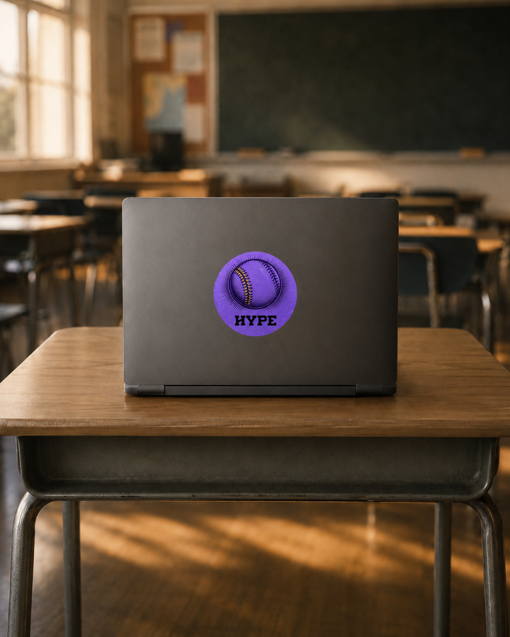 A dark gray laptop sits open on a wooden classroom desk, displaying a circular purple baseball sticker with the text &